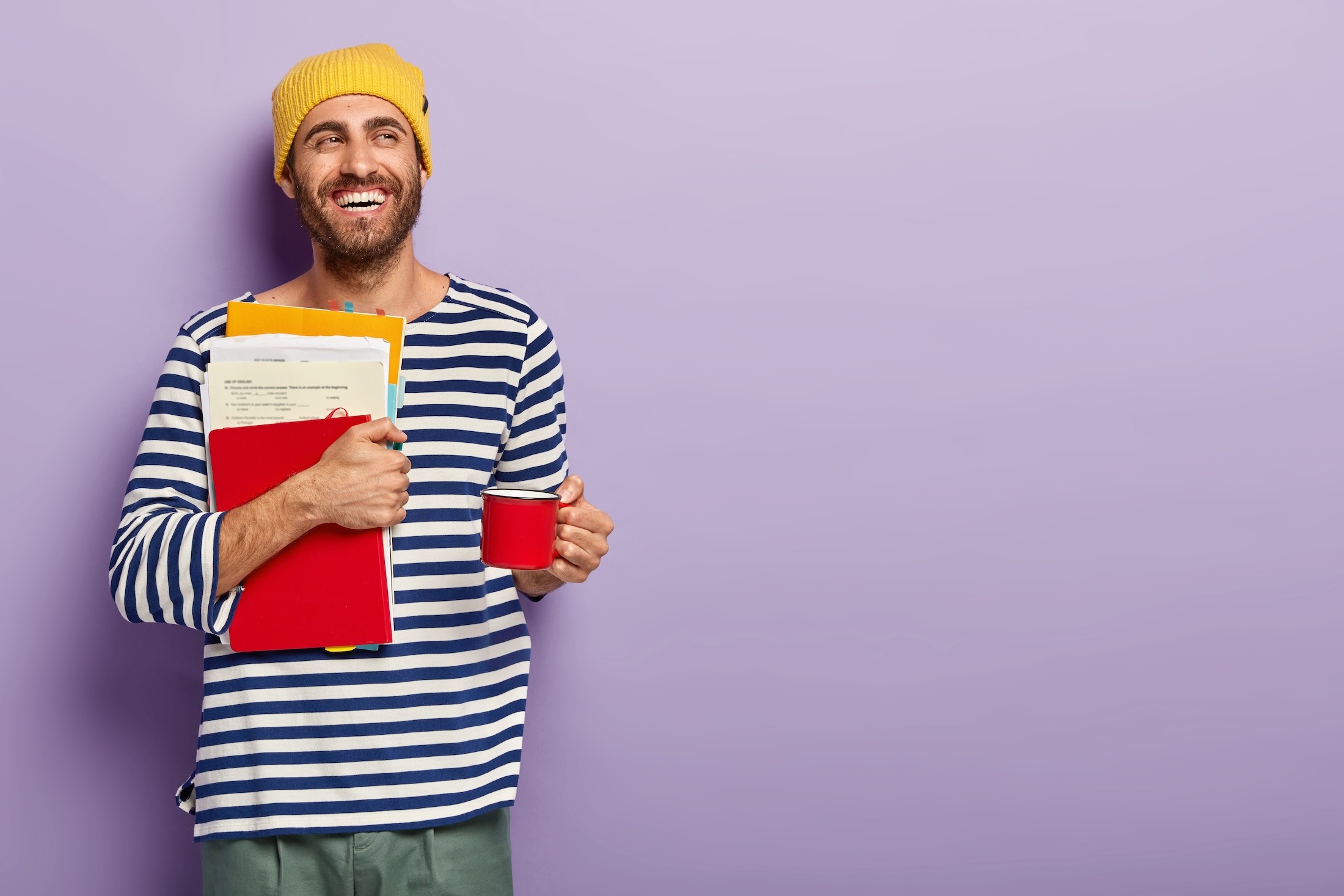 delighted-unshaven-young-man-student-holds-papers-red-notepad-holds-cup-with-hot-drink-has-coffee-break-being-good-mood-looks-aside-with-broad-smile-isolated-purple-background